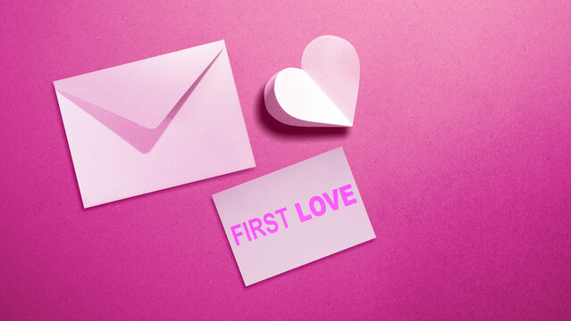 Flat lay of a pink love letter envelope, a 3D paper heart, and a card with the text 'First Love' on a textured pink background. Concept of First Love Day
