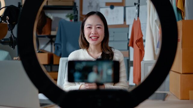 Young Asian woman fashion entrepreneur live streaming selling clothes online with smartphone ring light and laptop in home studio, representing small business owner and professional women.