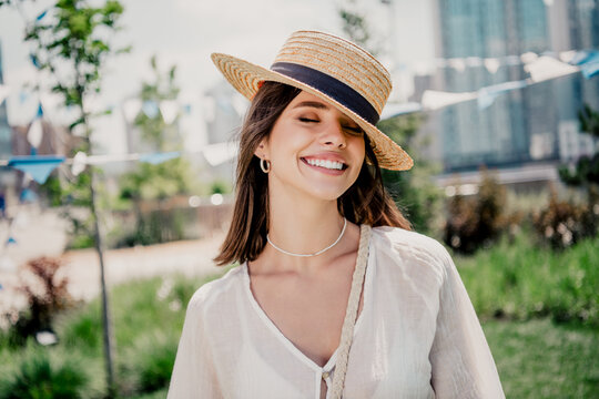 Smiling young woman in straw hat enjoying a sunny summer day outdoors in an urban park, casual white blouse and crossbody bag in a relaxed, joyful portrait