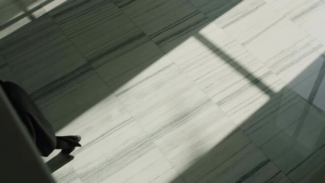 Overhead view of person walking across sunlit marble lobby beneath long window grid shadow, showing dark trousers, shoes, and coat hem in modern interior, with marble, lobby, atrium, walking detail