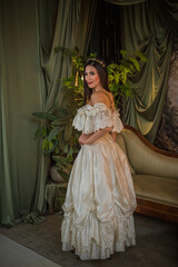 Fototapeta premium Elegant woman in a vintage ball gown sitting on a velvet sofa in a luxurious dark interior. Romantic gothic aesthetic portrait with crystal chandelier and antique atmosphere