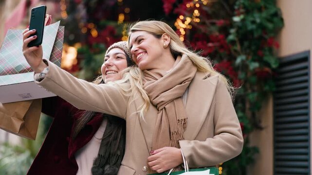 Video of two beautiful friends doing videocall with smartphone while walking on the city street with shopping bags.