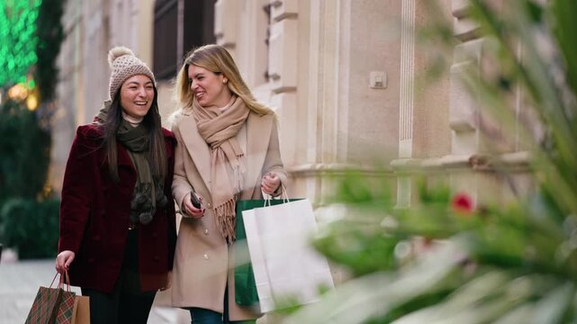 Video of two beautiful friends talking and having fun while walking on the city street with shopping bags.