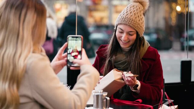 Video of one friend is taking a photo of the other with the Christmas gift she has received on a bar terrace