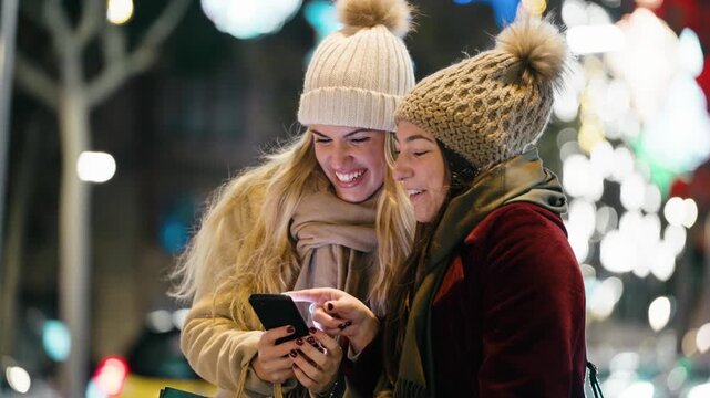 Video of two beautiful friends using smartphone while walking on the city street with shopping bag.