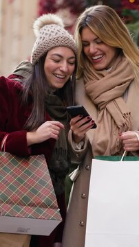 Video of two beautiful friends talking and using smartphone while walking on the city street with shopping bags.