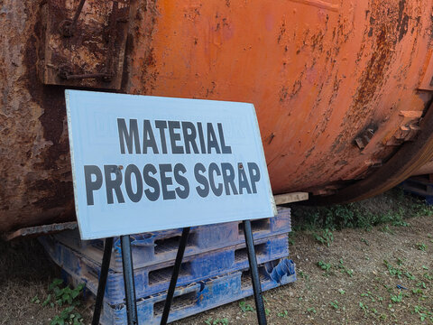 A warning sign indicating scrap material processing placed in front of a rusty industrial metal tank and plastic pallets. Ideal for industrial safety documentation, recycling concepts, manufacturing
