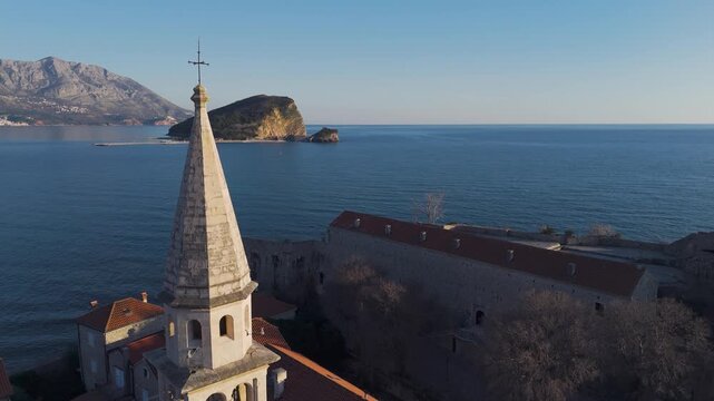 Cinematic aerial drone flyby close to the historic bell tower of Budva Old Town, revealing St. Nicholas island and a panoramic view of terracotta tiled roofs at sunset, Montenegro.