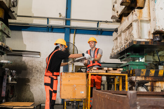 Coworkers exchanging stamped metal sheet part at industrial press station supporting efficient production and workplace safety inside a busy metalworking factory setting