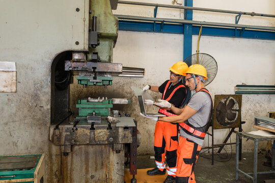 Two engineers reviewing laptop data beside large industrial power press machine