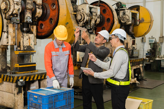 Senior manager inspecting metal bracket against light with engineer and press operator nearby highlighting skilled labor and industrial teamwork in the automotive parts manufacturing sector