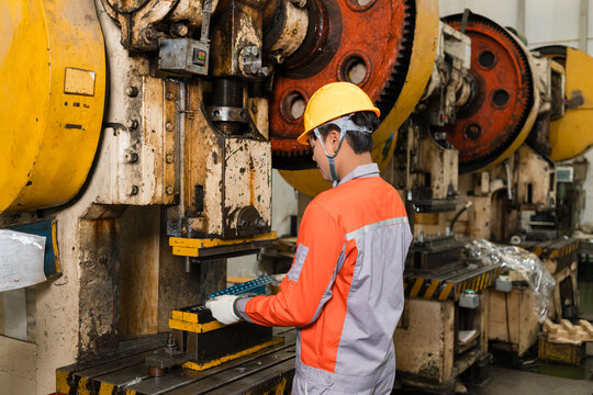 Male machine operator in a yellow hard hat holds a metal part up to check fit at a power press die