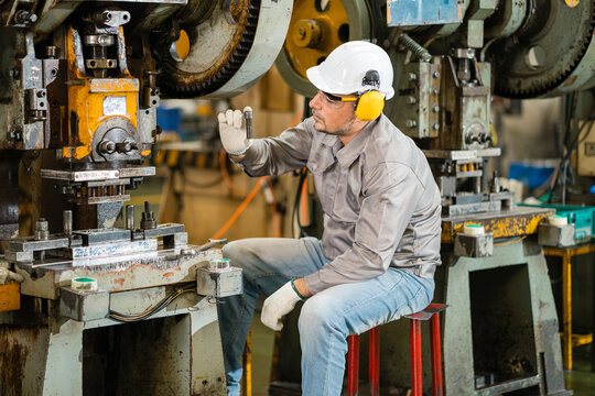 Male maintenance technician sitting on a stool between two power presses examining a steel bolt on the factory floor