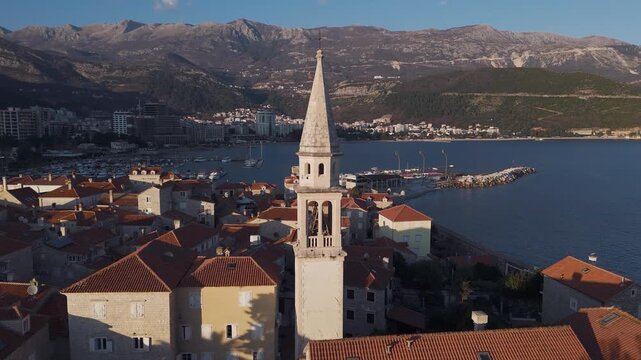 Cinematic aerial drone view flying directly towards the historic bell tower of Budva Old Town, with the sea, modern city and high mountains in the background, Montenegro.