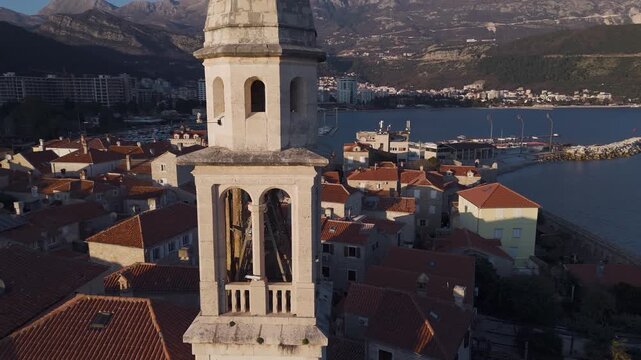 Close-up aerial drone orbit view around the historic bell tower of Budva Old Town, revealing terracotta roofs, new modern city and mountains at sunset, Montenegro.