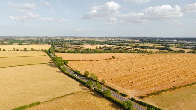 Round hay bales across harvested grain field. Straw bales scattered after wheat and barley harvest with cultivated farmland, country road and rural landscape. Livestock fodder production, post harvest