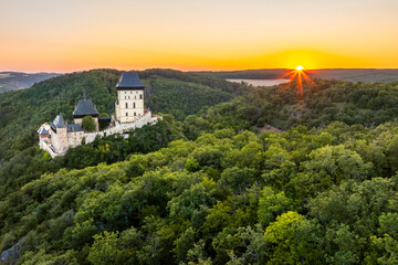 Fototapeta premium Aerial view of iconic gothic royal Karlstejn Castle surrounded by lush green forests at golden hour sunset, UNESCO candidate site, Central Bohemia, Czech Republic ( Hrad Karlštejn-Česká Republika )