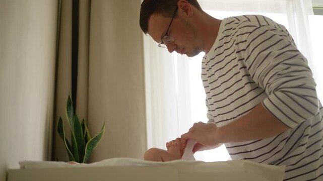 Young dad with glasses carefully puts a new disposable diaper on his infant son lying on a changing station, ensuring comfort and cleanliness as part of their daily childcare routine