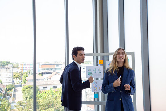 Two business professionals discussing marketing strategy and business analytics in front of a glass board with charts and sticky notes.Corporate colleagues collaborating on project planning,