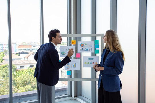 Two business professionals discussing marketing strategy and business analytics in front of a glass board with charts and sticky notes.Corporate colleagues collaborating on project planning,