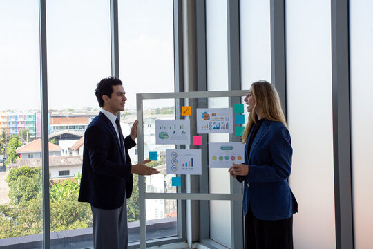 Two business professionals discussing marketing strategy and business analytics in front of a glass board with charts and sticky notes.Corporate colleagues collaborating on project planning,