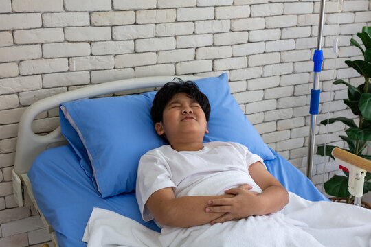 A sick boy lies in a hospital bed, one hand clutching his stomach, resting during treatment and recovery. This relates to healthcare, child care, illness, fever, patient care, and hospitals.