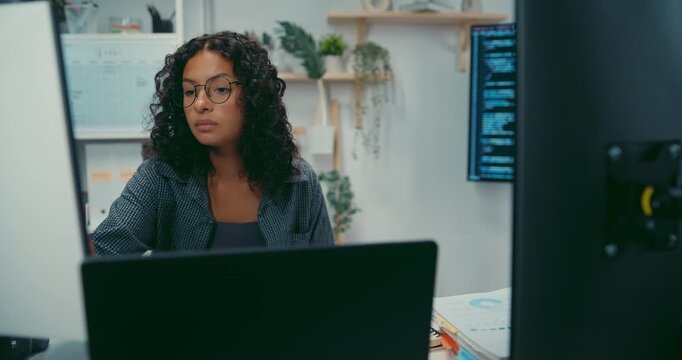 Young black female developer wear glasses writes complex software code at desk in office. Artificial intelligence machine learning, Secure computer network, Advanced cloud data analysis trend concept.