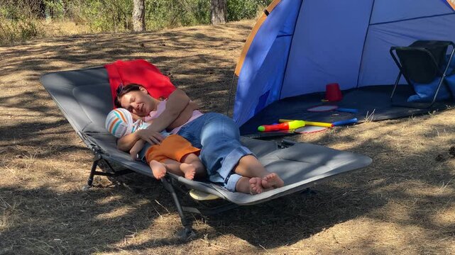 A mother gently hugs her small child while relaxing on a camping lounger. They rest together next to a blue tent in a sunny pine forest. This sweet cuddle feels tender and peaceful.