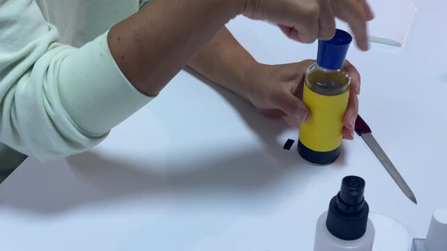 A woman's hands carefully open a small yellow cosmetic bottle on a white table. She works with calm focus and gentle precision. This quiet activity feels peaceful and satisfying.