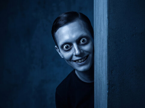 Creepy man with wide staring eyes and a sinister grin peeking from behind a wall in dark blue lighting perfect for horror thriller and mystery posters