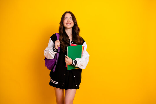 Smiling student with backpack and notebooks wearing a varsity jacket against a bright yellow background