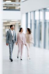 Fototapeta premium A focused businessman walks through a bright modern office corridor as blurred colleagues pass beside expansive windows. Motion blur, selective focus.