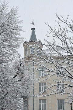 A detailed shot of a historic European building corner tower with a weather vane, covered in fresh snow and framed by snow-laden tree branches during a winter snowfall.