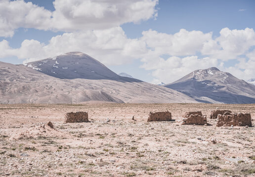 Ruins of ancient Pamir dwellings in the Tien Shan Mountains of Tajikistan on a high plateau with a mountain with cliffs and snow in the background. Landscape in sunny weather