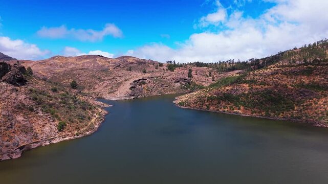 Aerial view moves over Las Ninas Dam, pine forests, and volcanic hills under blue sky