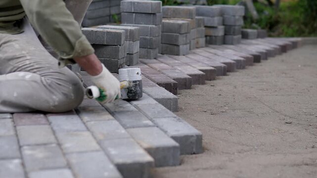 Worker kneeling on left side taps concrete paving block into place with mallet and gloved hands, forming straight row beside bare dirt path, stacked pavers, soft daylight, rough