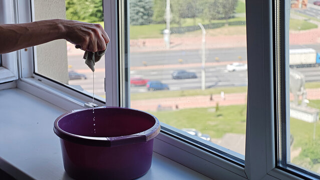 Hand wringing out a damp cloth into a purple basin, cleaning apartment window overlooking city street with cars