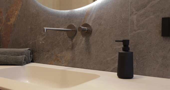 Backlit round mirror and wall mounted faucet above an integrated sink, camera glides left to right across dark stone cladding pale countertop folded towels and matte accessories