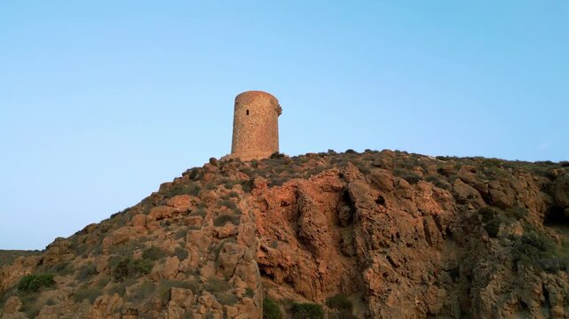 Ancient stone watchtower stands atop a rugged, sun-lit rocky hillside beneath a clear blue sky, evoking coastal Mediterranean heritage
