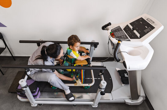 Cute young boy patient enjoys a session of physical rehabilitation, supported by a therapist using rehab walker while standing on a treadmill in a light-filled space