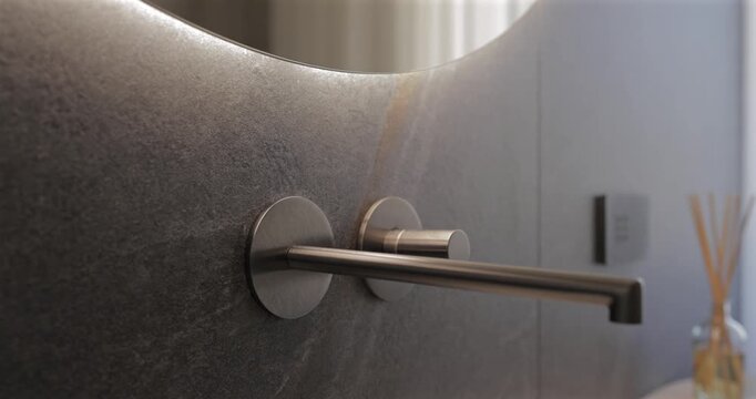 Sensor activated mirror glow above a brushed wall faucet in dark stone bathroom, static close video follows hand passing beneath the rounded edge as soft backlight awakens reshapes premium sink area