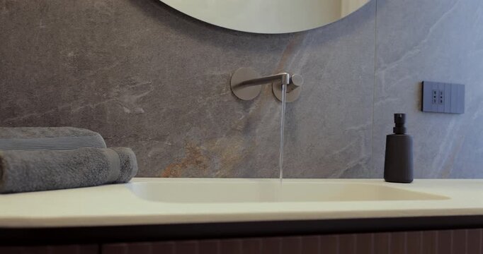 Bathroom interior with flowing water from wall faucet and curved wood vanity, left to right camera glide reveals the stone countertop round mirror folded towels black soap dispenser and reed diffuser
