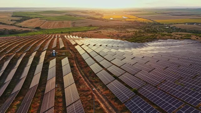 Solar panels are arranged in rows on a vast field in a rural location. The sun sets in the background, casting light over the landscape. Fields stretch beyond the solar installation.