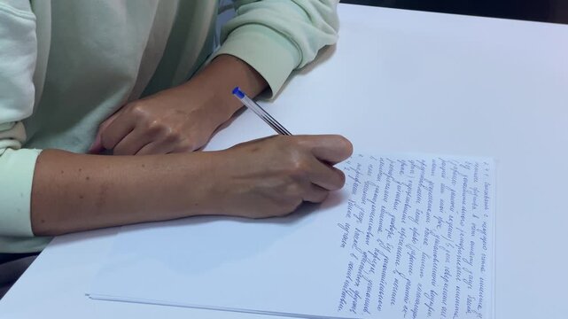 A student writes detailed cursive notes using a blue pen. Hands rest peacefully on a clean white desk in a quiet room. The tranquil mood reflects steady focus and calm diligence.