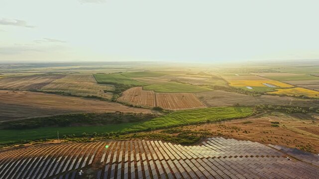The sun rises over a vast landscape with solar panels and farmland. Fields stretch under the sunlight as crops grow in rows. The scenery shows different shades of green and brown.