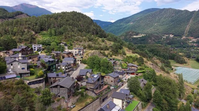 Aerial view of La Massana, Andorra, where traditional stone-and-slate homes cluster along a winding road beneath lush forested Pyrenean mountain slopes in summer