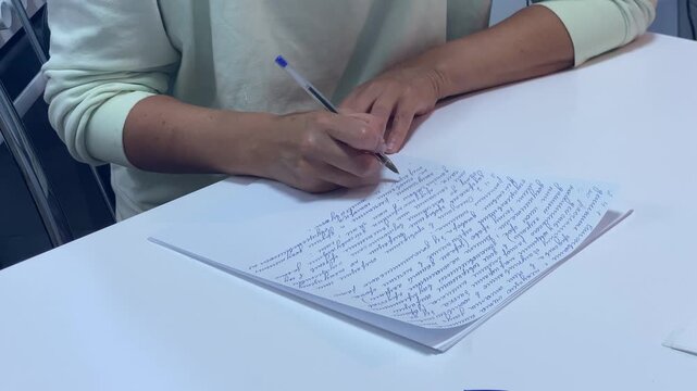 A student writes notes carefully on white paper with a pen. She sits comfortably at a clean white desk. Her steady focus creates a calm and peaceful mood.