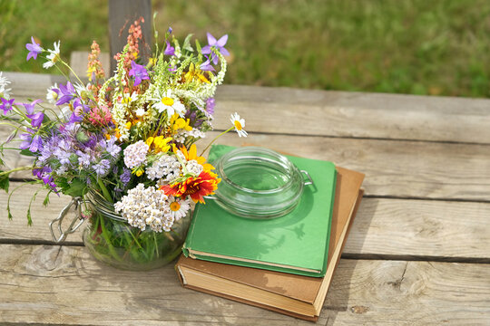 glass jar full of different colorful flowers and books on wooden bench in garden. summer season. Healthy plants harvest for preparation infusion, tea. Reading, knowledge botany, study of herbs