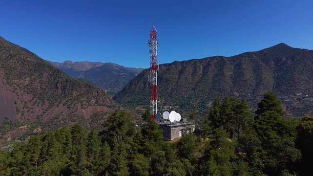 Red and white telecommunications tower with satellite dishes atop a stone building on a forested mountain ridge in Andorra, overlooking a valley town surrounded by steep slopes