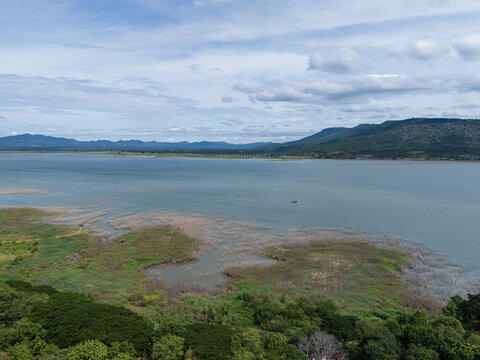 Drone aerial view of a large freshwater lake bordered by lush forest and distant mountains beneath expansive blue skies, showcasing natural landscape and tranquil scenery